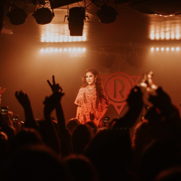 A performer in an orange outfit stands on stage, smiling under bright lights, as an enthusiastic audience raises their hands and takes photos in a dark concert venue.