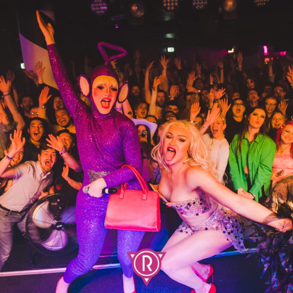 Two drag performers pose energetically on stage in front of a cheering crowd. One is dressed as a sparkling purple Teletubby with a pink purse, and the other is in a sparkling, revealing outfit with platinum blonde hair.