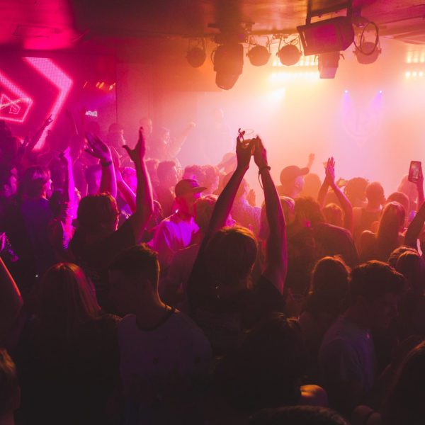 A crowded nightclub with people dancing under colorful neon and yellow lights. Many have their hands raised, and a lively, energetic atmosphere fills the room.