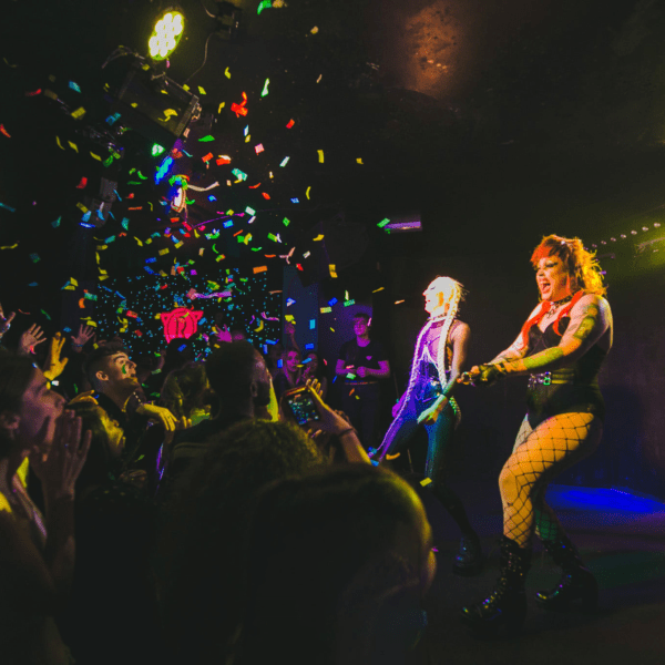 Two drag performers in colorful outfits entertain a cheering crowd on a dark stage, with bright lights and confetti filling the air, creating a lively and festive atmosphere.