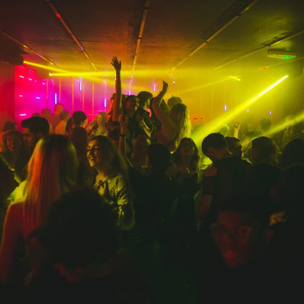 A crowded nightclub with people dancing under colorful lights, including purple and yellow beams, creating a lively and energetic atmosphere. Some people have their arms raised, enjoying the music and ambiance.