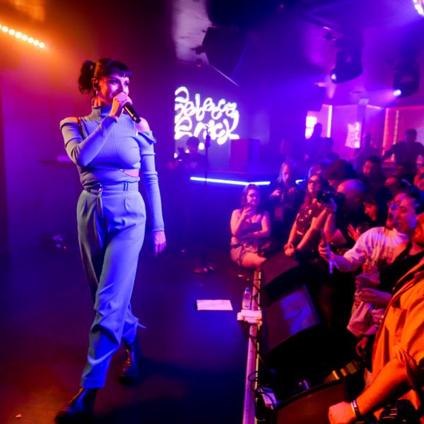 A female singer in a blue outfit performs on stage under colorful lights, holding a microphone, while a lively crowd watches and takes photos in a packed nightclub.