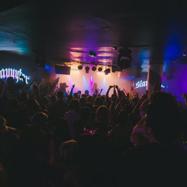 A large crowd enjoys a concert in a dimly lit indoor venue, with colorful stage lights illuminating the performer and raised hands visible throughout the audience.
